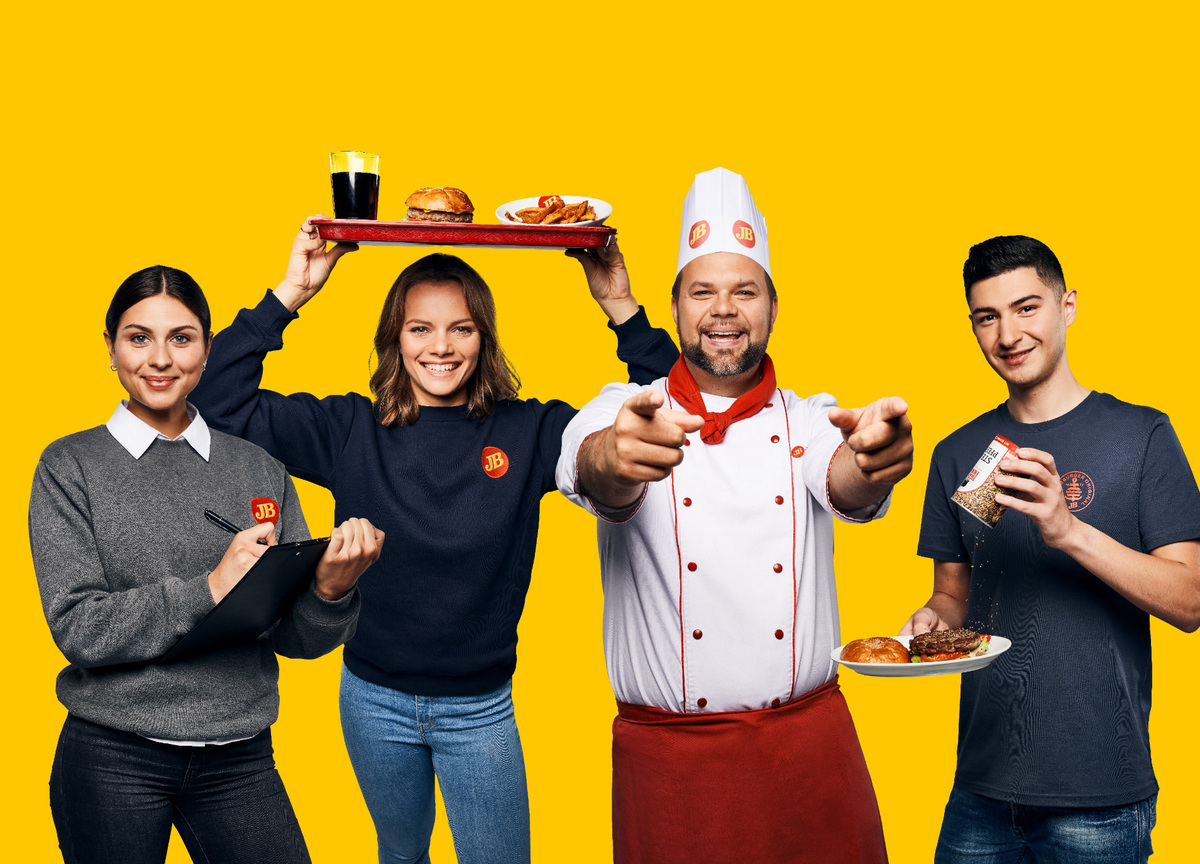 Jim-Block-Team in Uniformen vor gelbem Hintergrund, fröhlich und einladend, mit Burgern und Getränken.