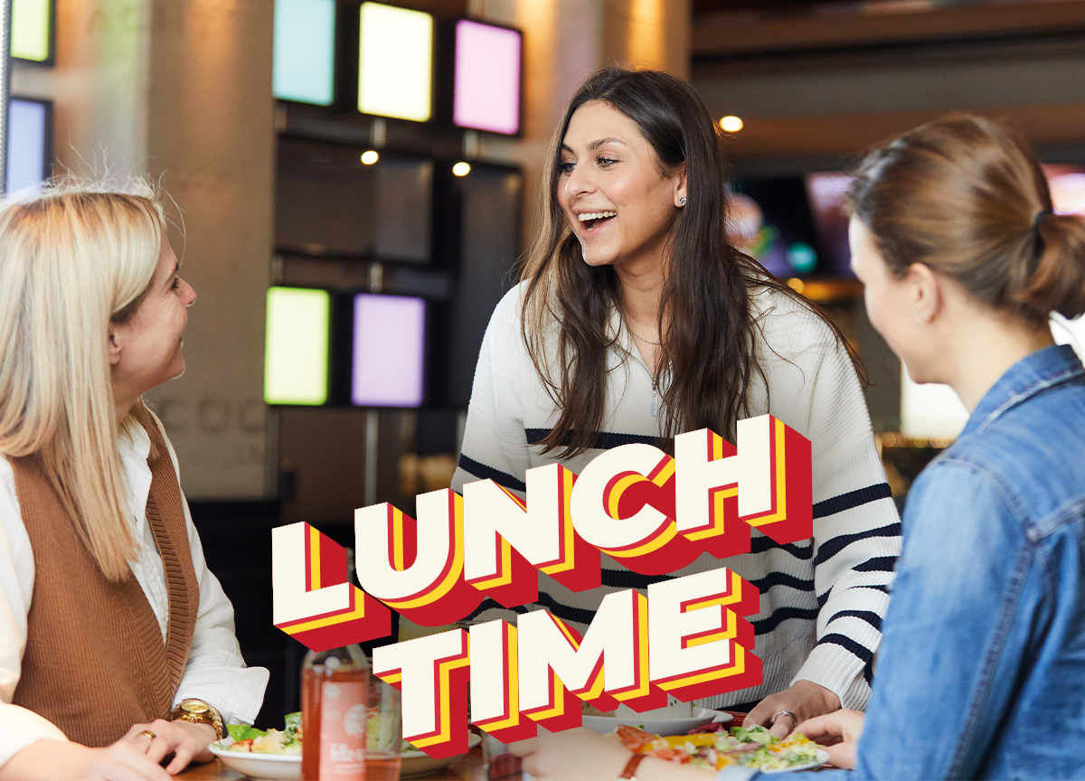 Jim Block Freunde essen Lunch Time Drei junge Frauen essen gemeinsam im Jim Block Restaurant und lachen – Lunch Time Schriftzug im Vordergrund