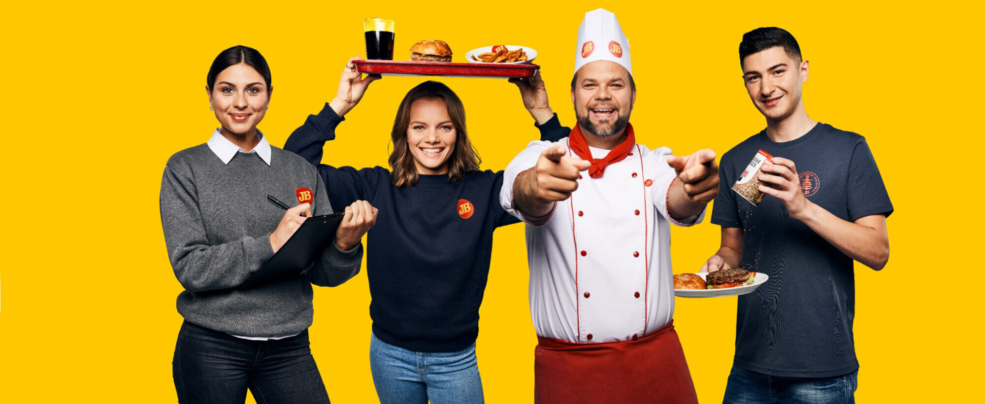 Vier bei Jim Block Arbeitende mit Essen stehen vor gelbem Hintergrund und lächeln