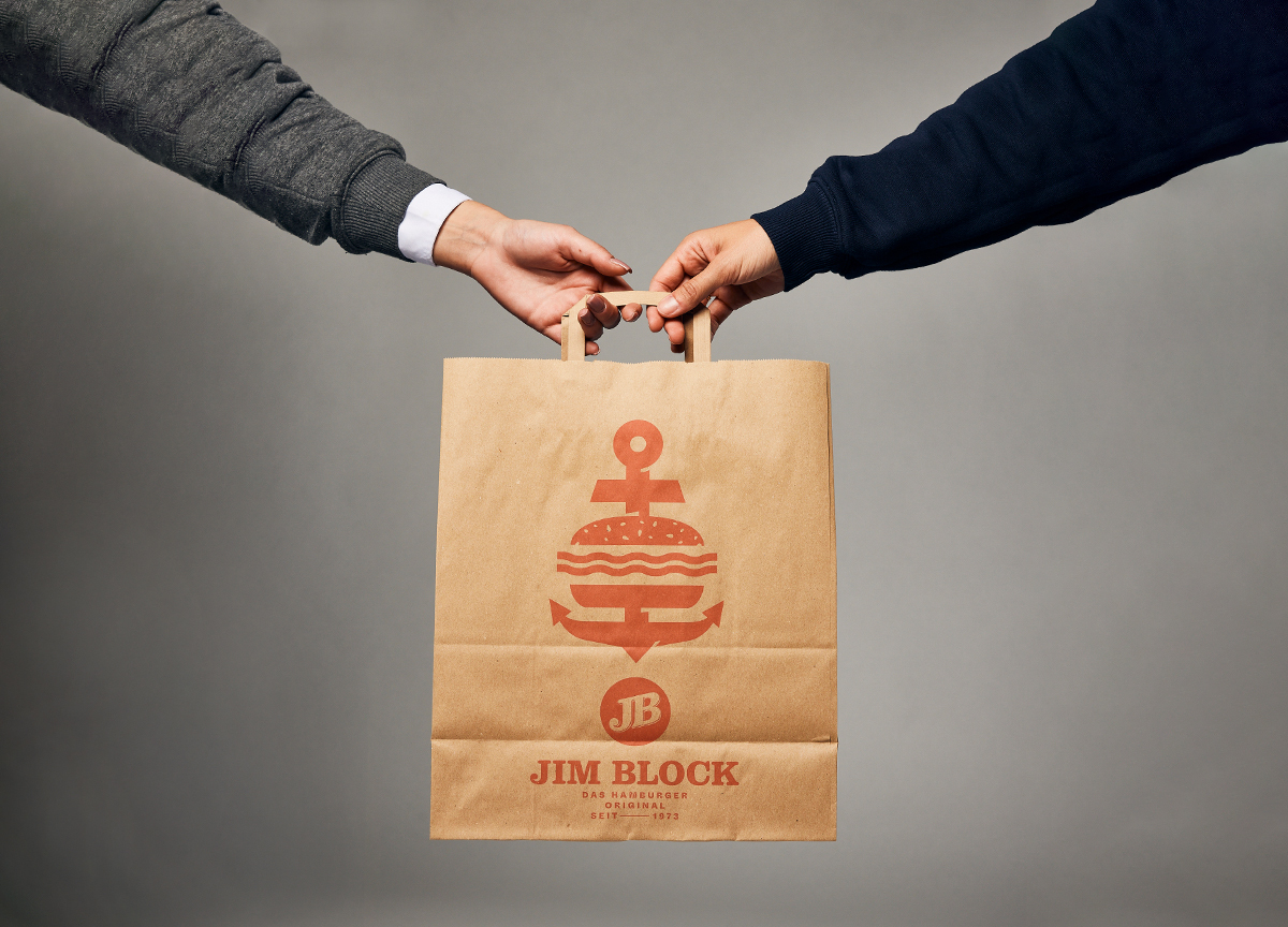 JB takeaway Tüte Zwei Hände übergeben sich eine braune Papiertüte mit Jim-Block-Logo.