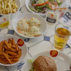 Auf dem Tisch sind mehrere Teller mit Essen angeordnet. Ein Teller enthält einen Hamburger mit Salat, daneben liegen Pommes frites und Süßkartoffelpommes. Ein weiterer Teller bietet einen gemischten Salat, eine Portion Krautsalat und ein Getränk in einem Glas.